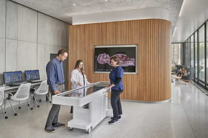 Three people stand around an Anatomage Table in the middle of the Countway Makerspace, with a large digital monitor on the wall.